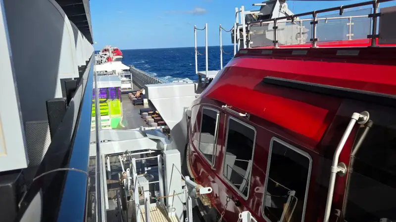 View from the side of a Virgin Voyages cruise ship, showing the ocean, ship structure, and emergency lifeboat.