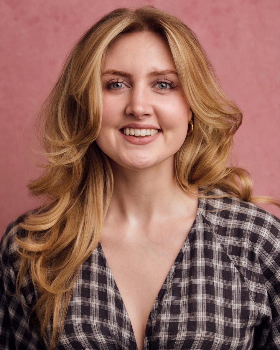 Smiling woman with long blonde hair in a plaid shirt against a pink background