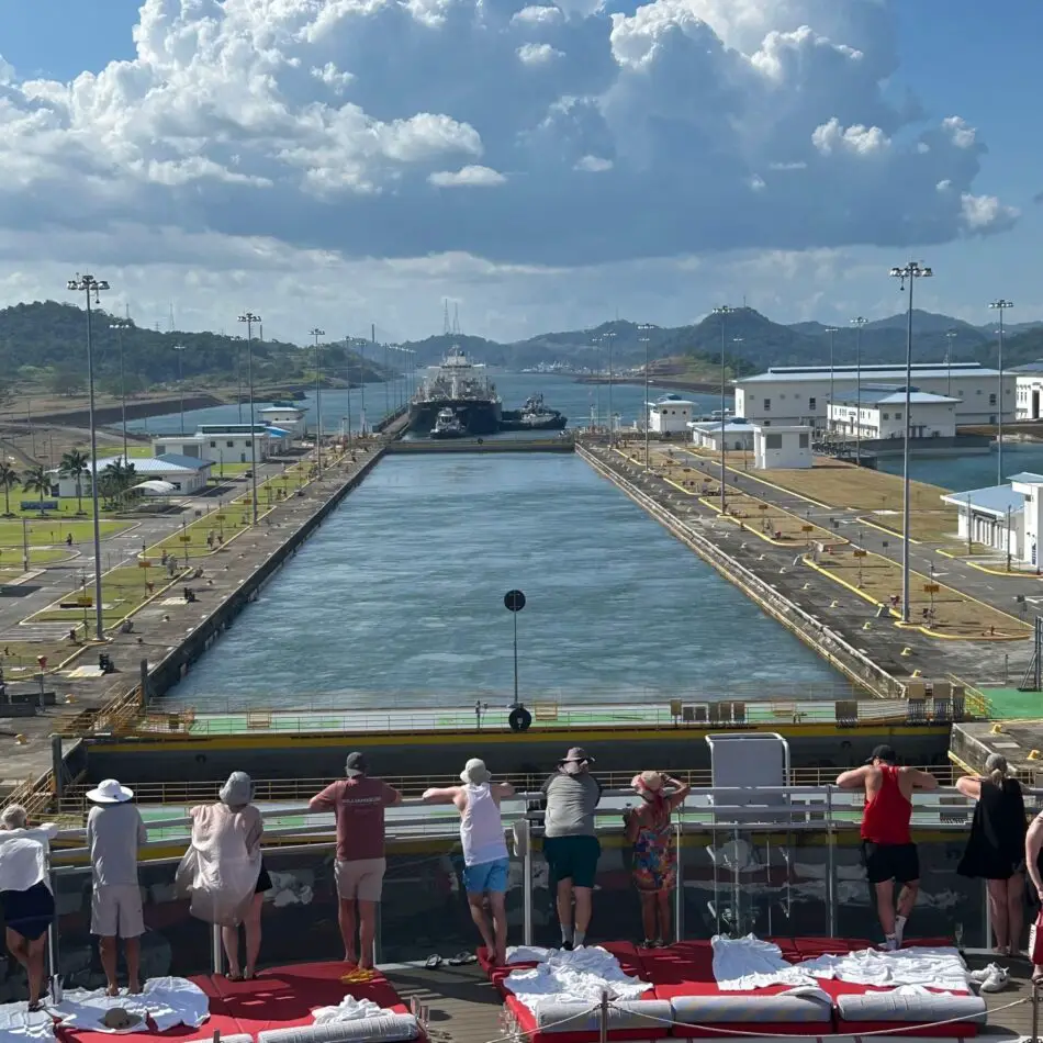 Passengers on a cruise ship deck watch a canal lock and distant vessel