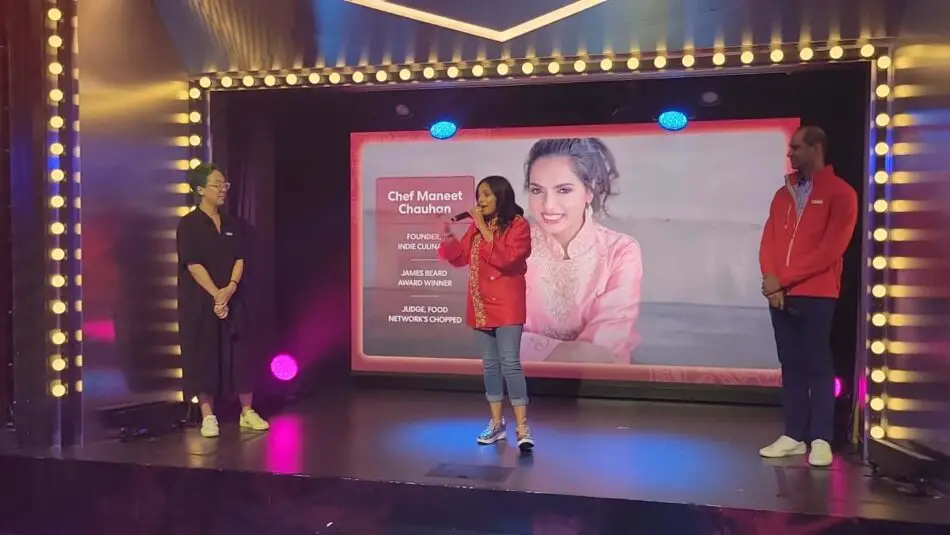 Three presenters on stage with a large screen displaying a smiling chef