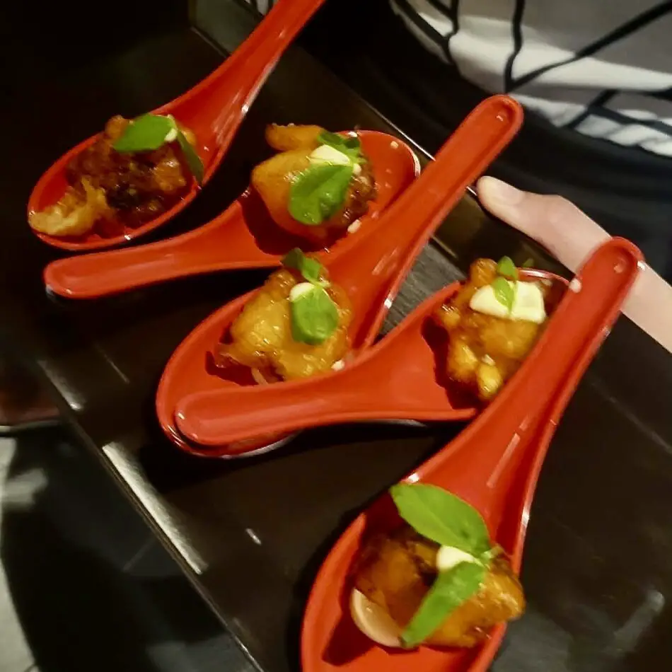 Four red ceramic spoons hold crispy fried bites topped with fresh basil leaves on a dark table
