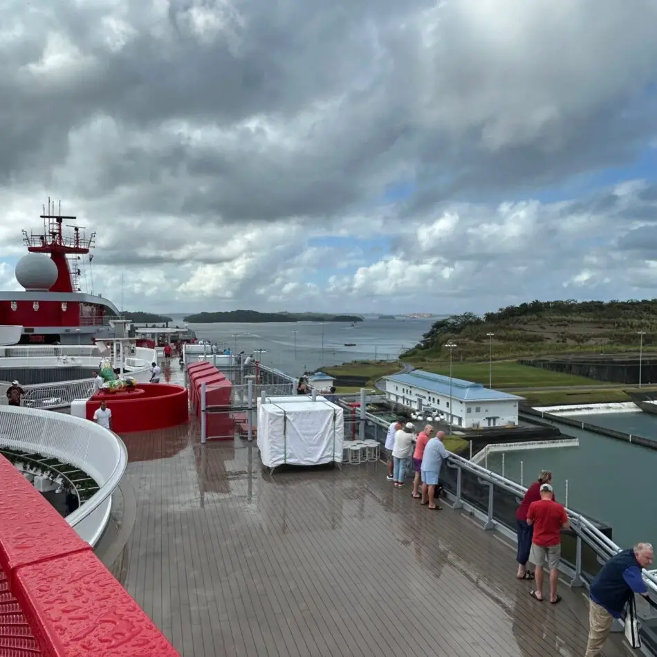 Deck of a Virgin Voyages cruise ship with passengers looking over the railing toward the harbor