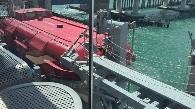 View from a Virgin Voyages cruise ship balcony, showing a city skyline and turquoise water with a red lifeboat below.