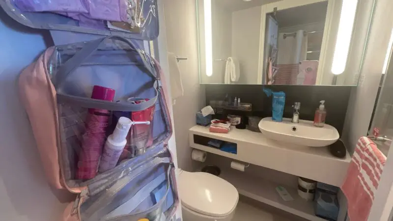 Virgin Voyages cruise ship cabin bathroom with a wall-mounted toiletry organizer and a sink surrounded by toiletries. The mirror has built-in lighting, and towels are visible near the sink and toilet.