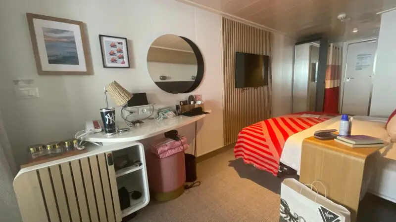 Virgin Voyages cruise ship cabin featuring a bed with a red and white striped blanket, a wall-mounted TV, a desk with a lamp, round mirror, and artwork on the wall.