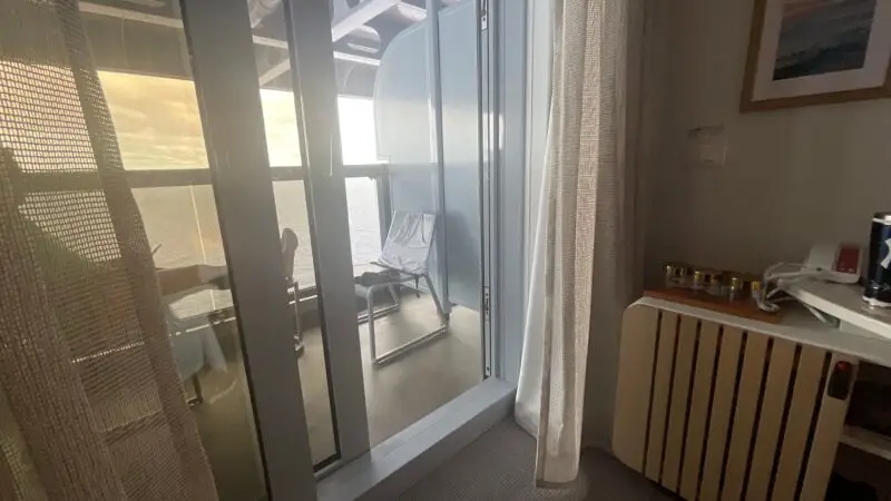 Cruise ship cabin with an open sliding glass door leading to a balcony. The balcony has two chairs and overlooks the ocean.