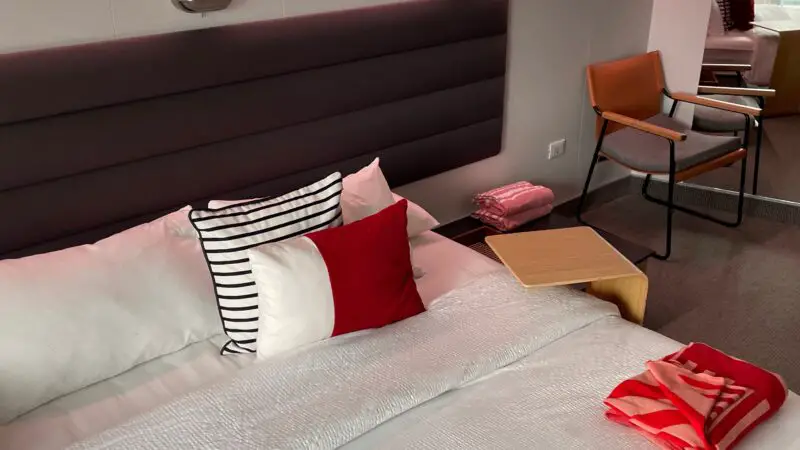 Virgin Voyages cruise ship cabin with a bed, modern lighting, a chair, and a small table.