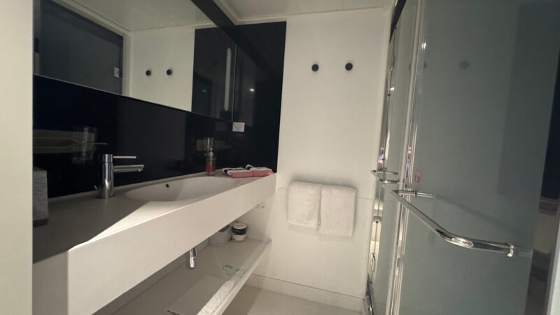 Virgin Voyages cruise ship cabin bathroom featuring a modern sink, large mirror, and glass shower door. Towels hang neatly on the wall.
