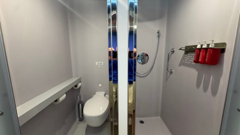 Bathroom of a Virgin Voyages cruise ship cabin, featuring a toilet, a shower area with a handheld showerhead, and three wall-mounted toiletry dispensers.