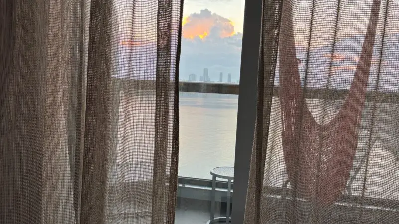Cabin view from a Virgin Voyages cruise ship showing sheer curtains, a balcony with a hammock and table, and a sunset over the ocean.