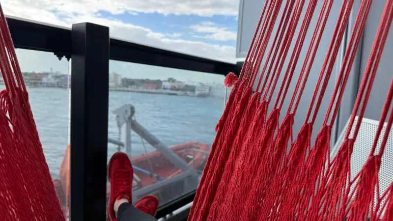 Passenger relaxing in a red hammock on a Virgin Voyages stateroom balcony, with a view of the ocean and distant coastline.
