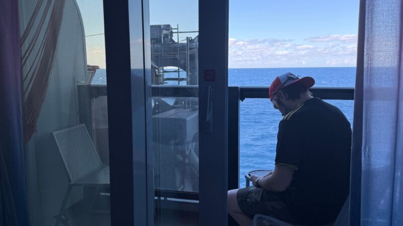 A Virgin Voyages cruise ship cabin with a glass door leading to a balcony. A person is seated on the balcony overlooking the ocean.