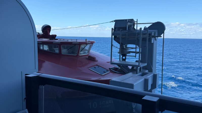 View from a Virgin Voyages cruise ship balcony showing a red lifeboat outside, with a metal chair in the foreground and blue ocean in the background.