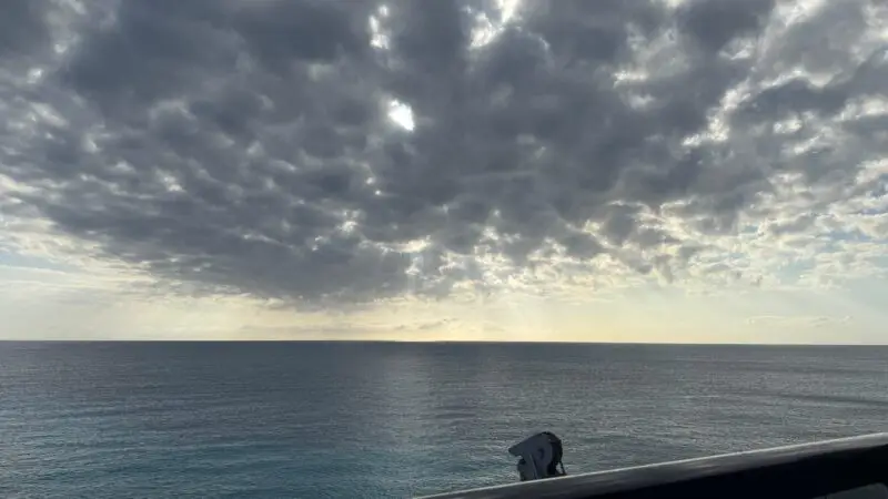 View from the balcony of a Virgin Voyages cruise ship cabin, showing a vast ocean under a cloudy sky with sun rays peeking through.