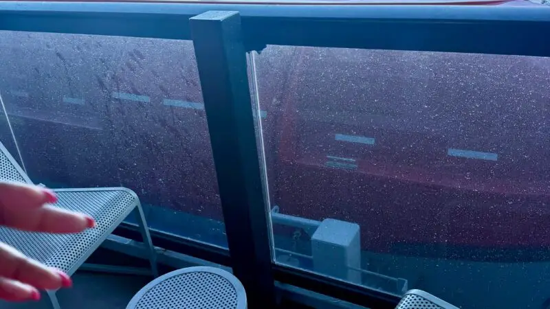 Balcony of a Virgin Voyages cruise ship cabin, featuring metal chairs and a small table, with an ocean view and coastline in the background.