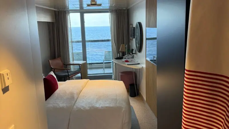 Cruise ship cabin with a bed, ocean view balcony, seating area, and desk.