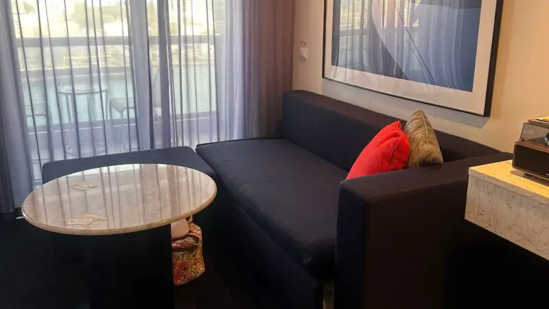 Virgin Voyages cruise ship cabin with a sofa, marble table, and large window with sheer curtains. Artwork hangs on the wall.