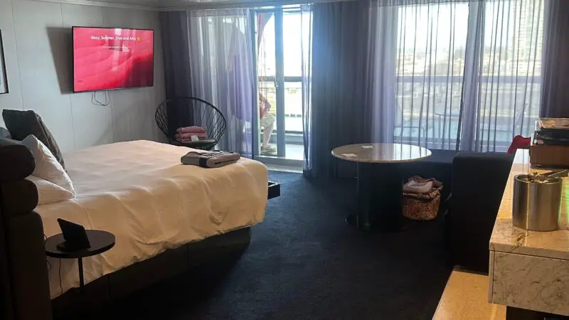 Virgin Voyages cruise ship cabin with a bed, TV, chair, table, and balcony access through glass doors.