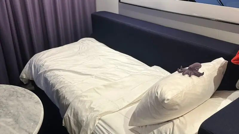 Virgin Voyages cruise ship cabin with a neatly made bed, a marble table, and a nautical-themed artwork on the wall.