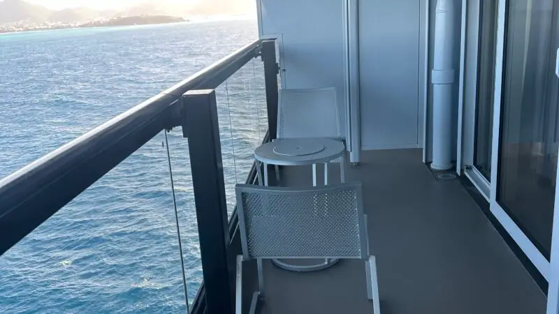 Balcony of a Virgin Voyages cruise ship cabin with two white chairs and a small round table overlooking the ocean and distant mountains.