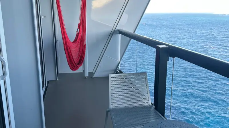 A balcony on a Virgin Voyages cruise ship cabin with a red hammock, metal chair, and round table, overlooking the ocean.