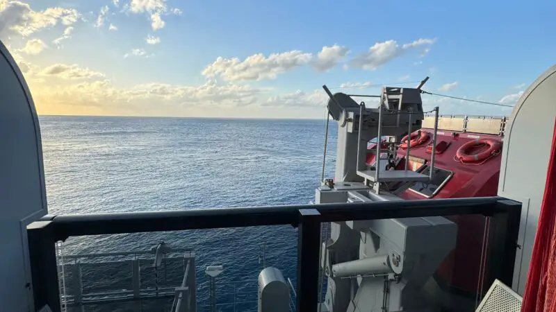 Balcony of a Virgin Voyages cruise ship cabin with two chairs and a small table, overlooking the ocean under a blue sky with clouds.
