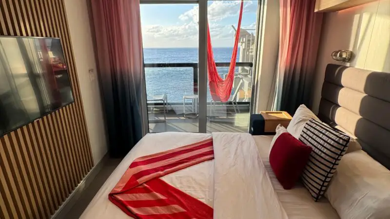 Virgin Voyages cruise ship cabin with a bed, colorful bedding, wall-mounted TV, and balcony view of the ocean with a red hammock.