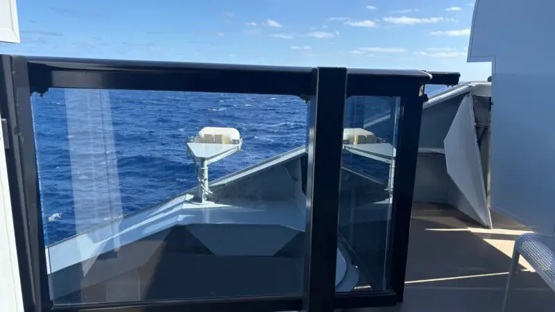 View from a Virgin Voyages cruise ship balcony with glass railing, overlooking the ocean and a clear blue sky.