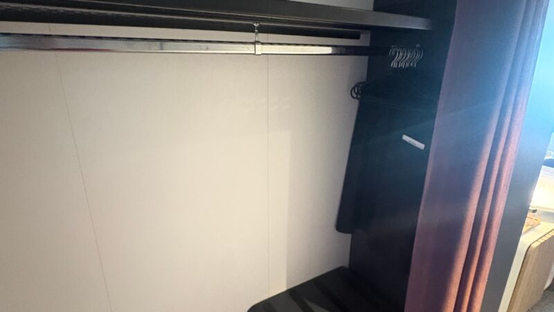 Interior of a Virgin Voyages cruise ship cabin closet, featuring a hanging rod with hangers, a shelf, and a small storage unit.