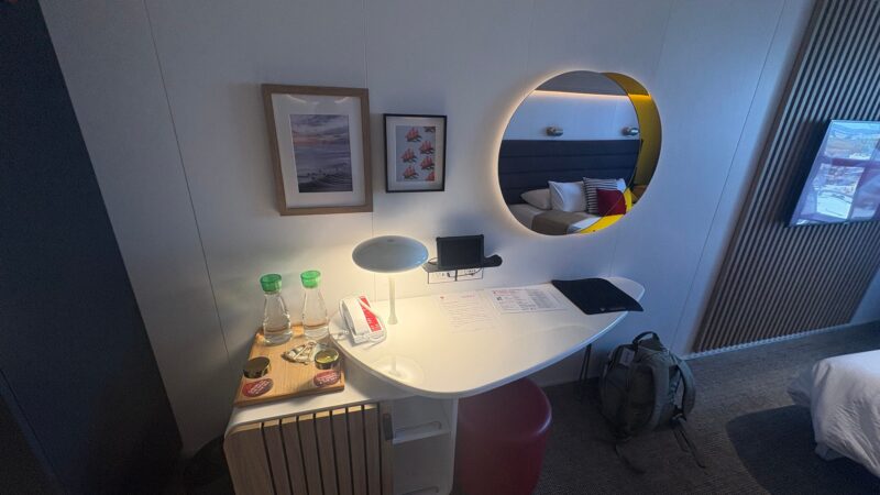 Virgin Voyages cruise ship cabin featuring a desk with a lamp, water bottles, artwork on the wall, a circular mirror, and a bed visible in the reflection. A backpack is on the floor.