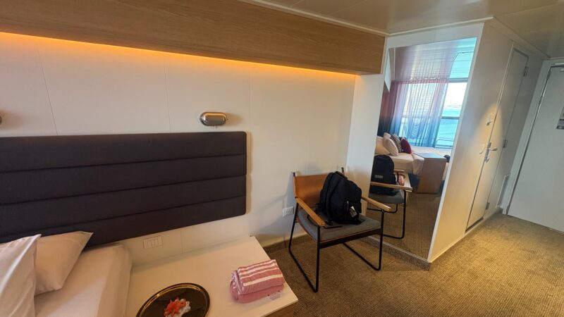 Virgin Voyages cruise ship cabin with a bed, soft lighting, a tray with towels, a chair with a bag, and a window with a view outside.