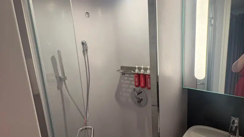Virgin Voyages cruise ship cabin bathroom featuring a modern shower with a glass door, mounted showerhead, and a sleek sink with an illuminated mirror.