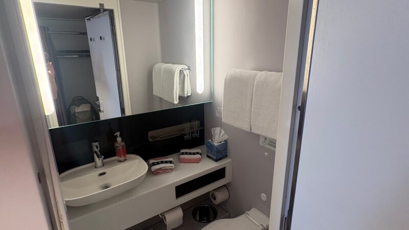 Bathroom in a Virgin Voyages cruise ship cabin, featuring a sink, toilet, towels, and toiletries on a counter.