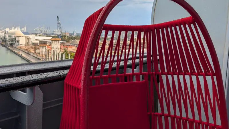 A Virgin Voyages cruise ship balcony with a red hanging chair, overlooking a port and cloudy sky.