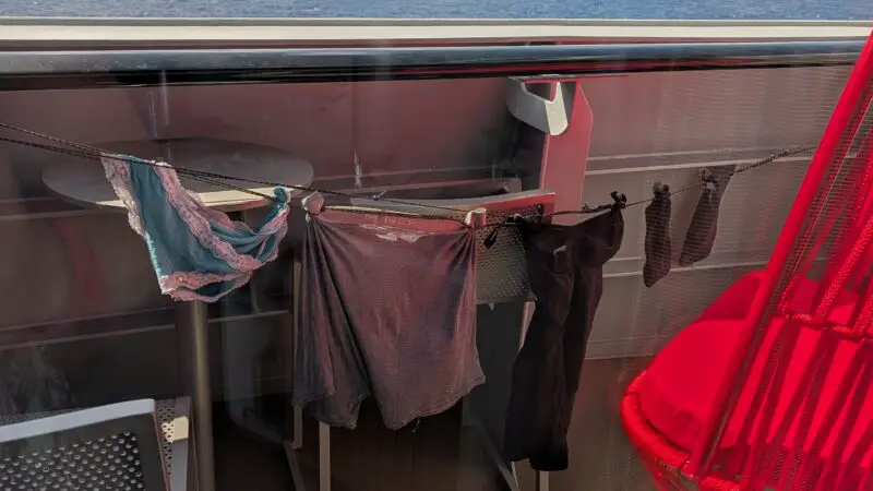 Balcony of a Virgin Voyages cruise ship cabin, featuring a red hammock chair, table, chairs, and clothes hanging to dry; ocean and sky visible in the background.
