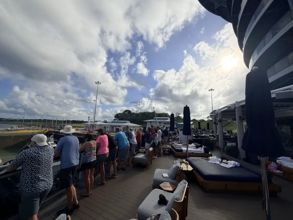 Passengers stand at the railing on a Virgin Voyages cruise ship deck, with outdoor lounge furniture