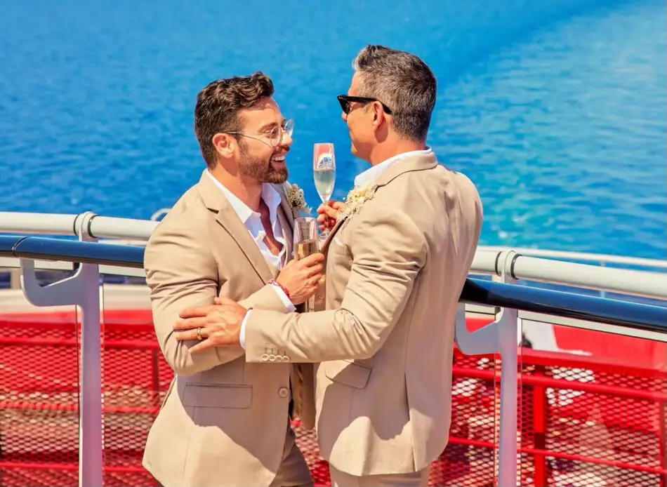 Two men in beige suits toasting champagne on a Virgin Voyages cruise ship deck