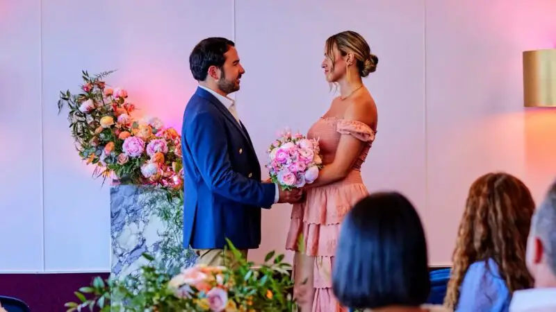 Couple in formal attire exchange wedding vows with pink bouquets aboard a Virgin Voyages cruise ship