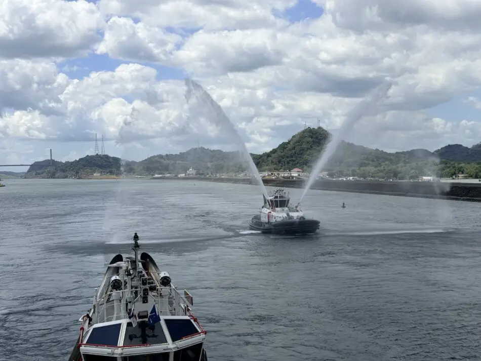 Two tugboats spray water from their cannons in a harbor with a hillside town in the background