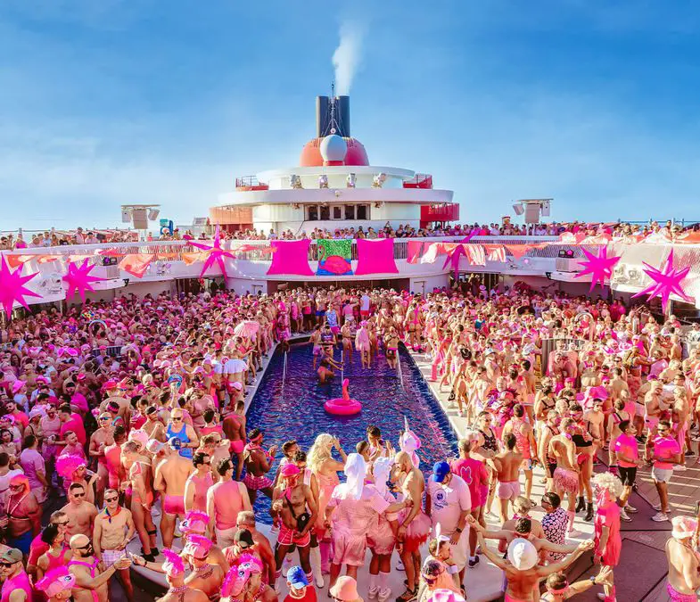 A large crowd dressed in pink surrounds a pool on a cruise ship deck under a clear blue sky.