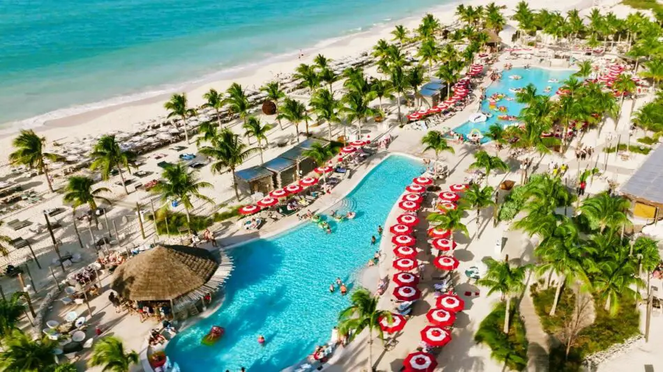 Aerial view of a tropical resort with swimming pools, red and white umbrellas, and palm trees near the beach.