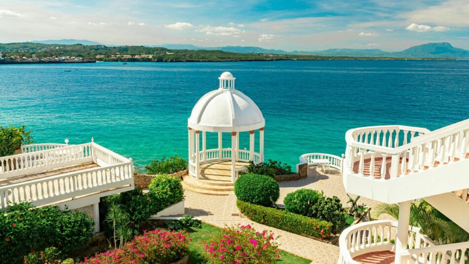 A white gazebo overlooks a turquoise ocean with surrounding tropical gardens and white staircases.