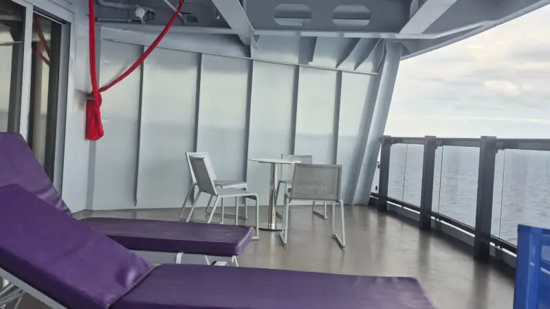 Balcony of a Virgin Voyages cruise ship cabin with purple lounge chairs, a table with chairs, a red hammock, and a view of the ocean.