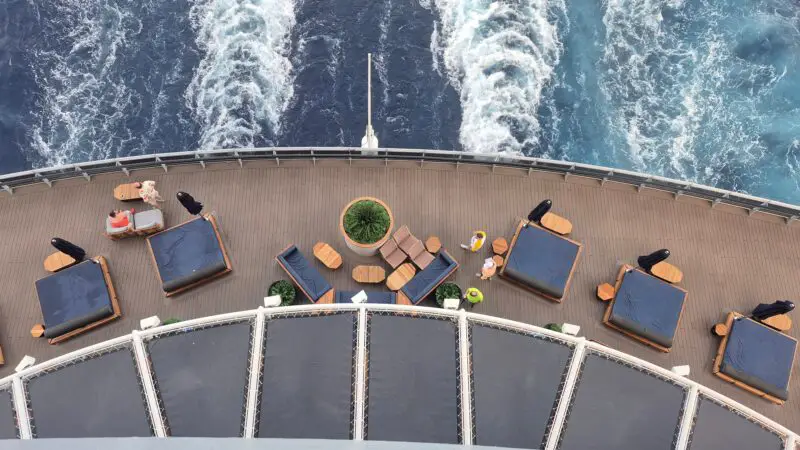 View from a Virgin Voyages cruise ship balcony, overlooking a deck with loungers and tables, with ocean waves trailing behind the ship.