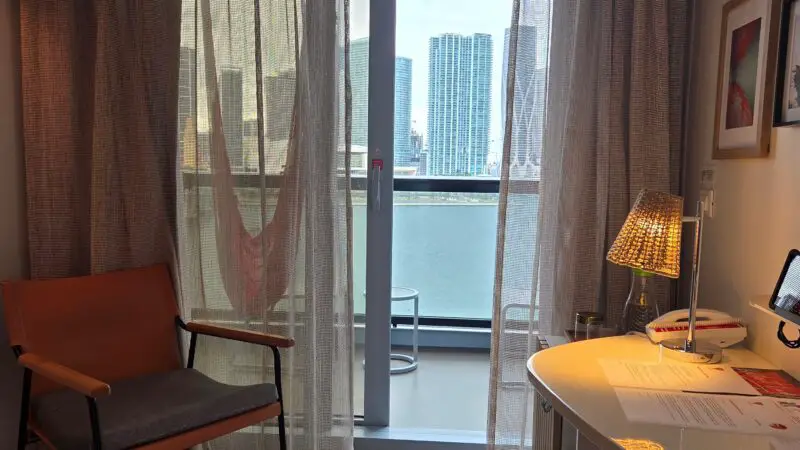Virgin Voyages cruise ship cabin with a balcony view of skyscrapers. The cabin features a chair, desk with papers, and floor-to-ceiling curtains.