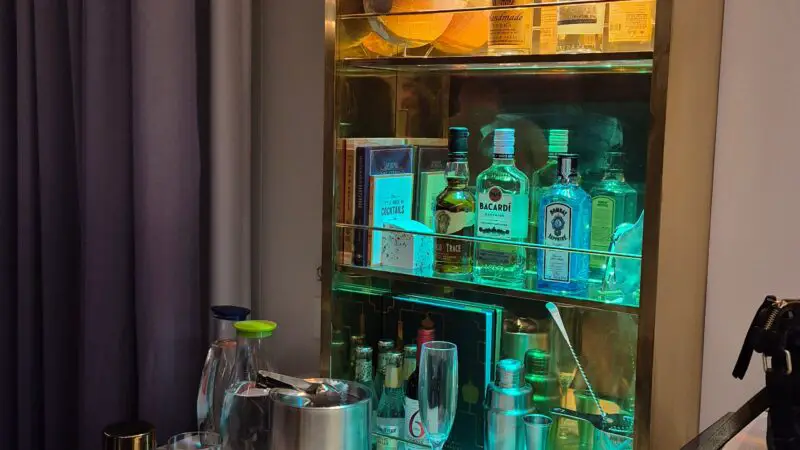 Virgin Voyages cruise ship cabin minibar with a marble countertop, liquor bottles, glasses, and cocktail accessories under soft lighting.