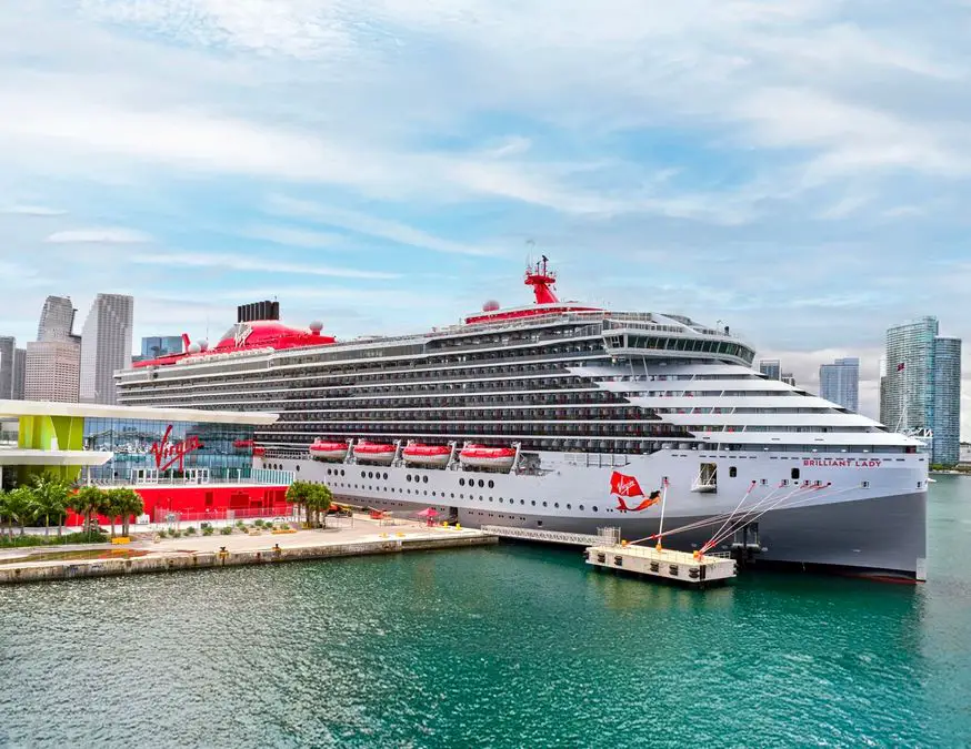 A large cruise ship docked at a port with a city skyline in the background.