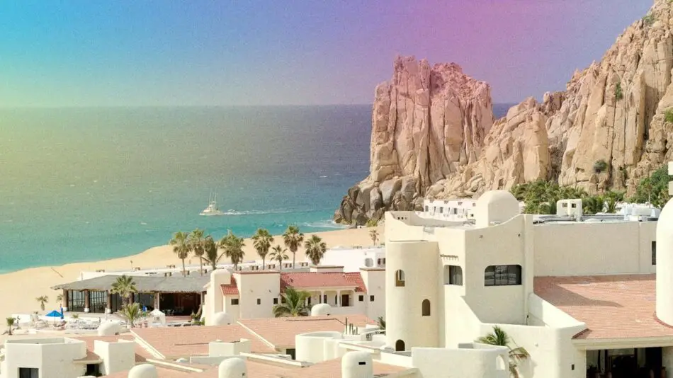 Beachfront view of white buildings, palm trees, blue ocean, and rocky cliffs under a colorful, gradient sky.