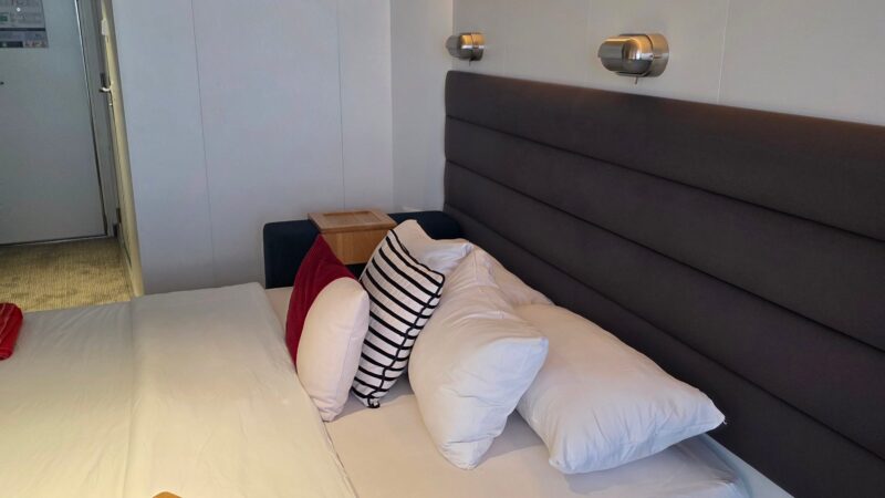 Cruise ship cabin with a bed, wooden side table, textured headboard, and accent lighting.