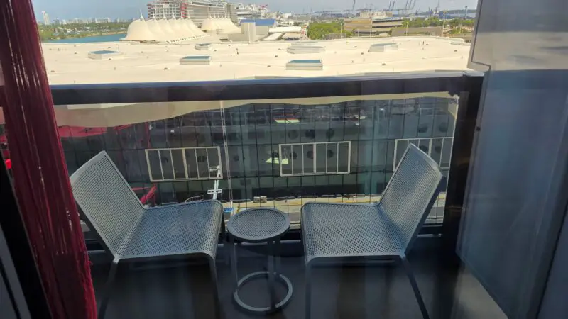 Cruise ship cabin balcony with two metal chairs and a small table overlooking a port with docked ships.